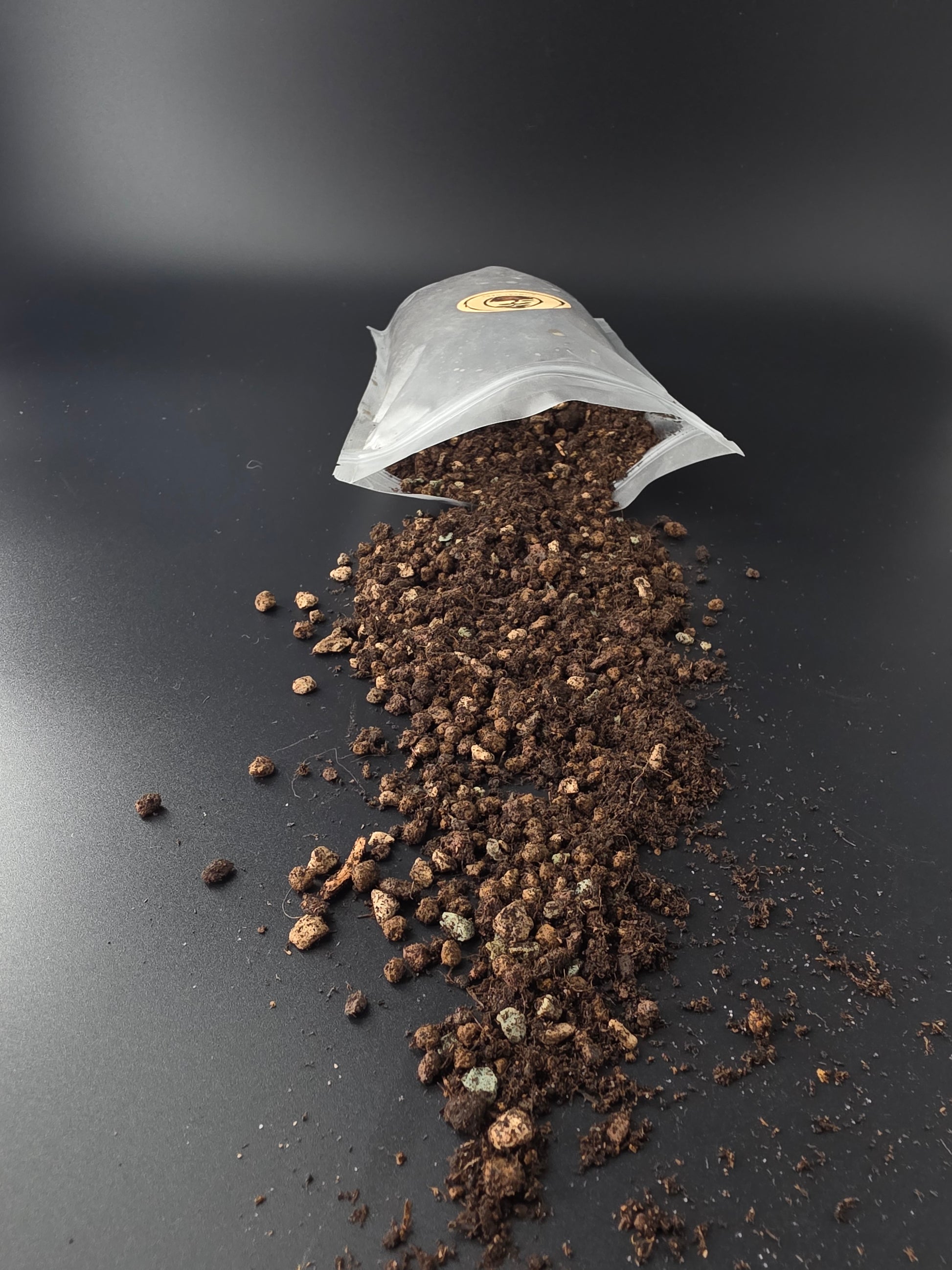 Open pouch on its side with dark, crumbly soil spread in a wide fan across the dark surface, fine dust scattered around.