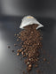Open pouch on its side with dark, crumbly soil spread in a wide fan across the dark surface, fine dust scattered around.