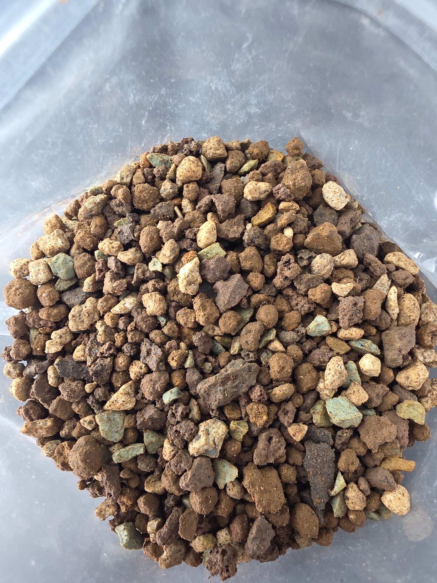 Close-up of chunky granules in tan, brown and pale grey-green piled on the crinkled clear plastic of the bag.