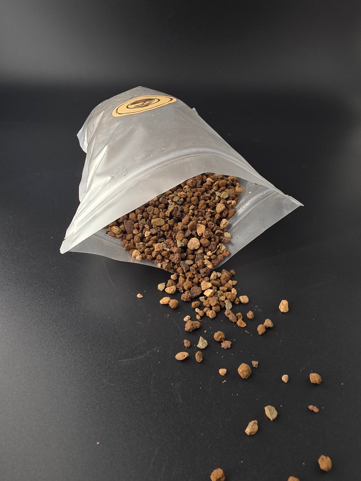 Pouch tipped on its side pouring coarse granules to the upper right, tan and dark grains scattered over the dark surface.