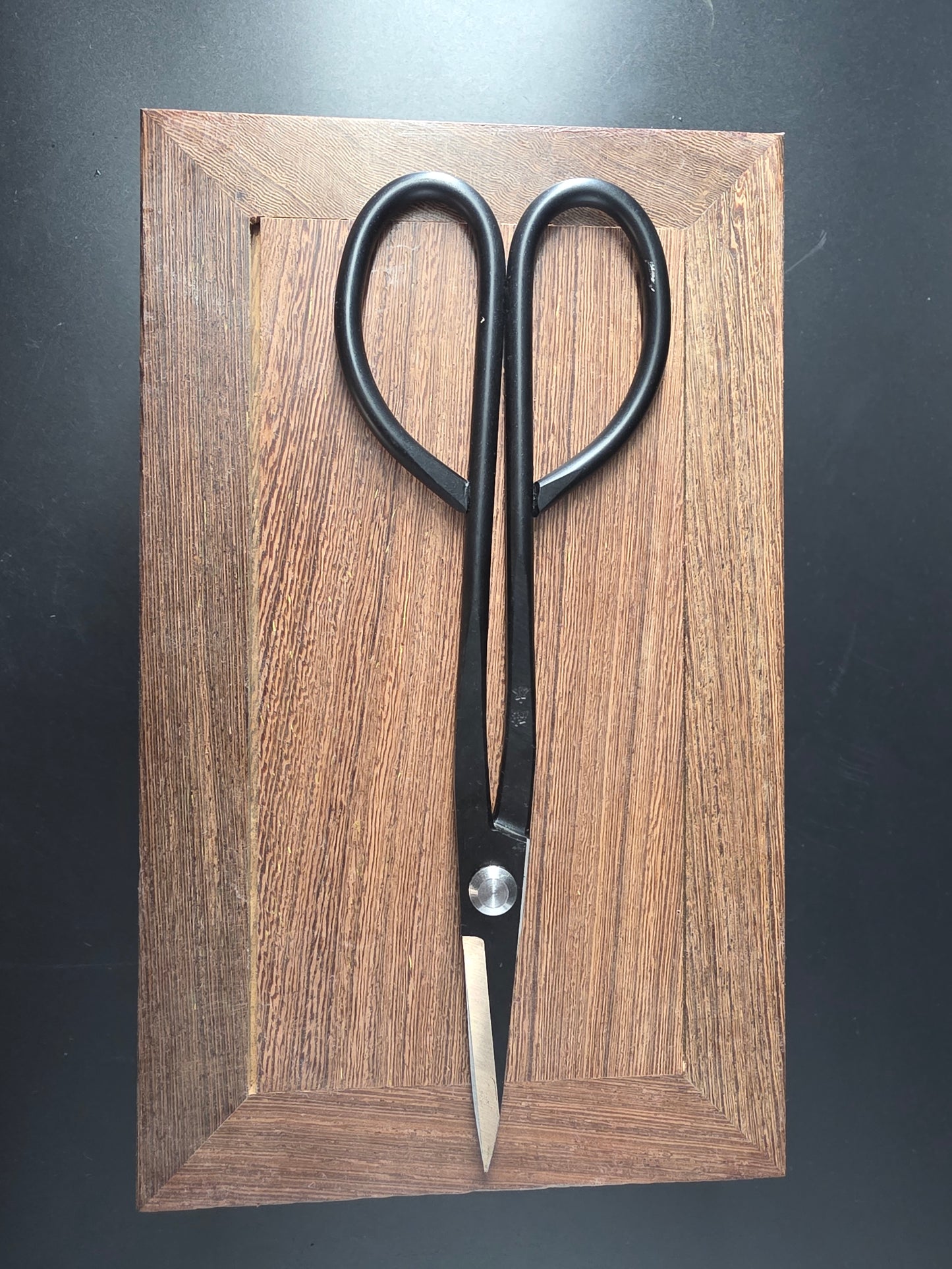 Black bonsai pruning scissors with loop handles and long narrow pointed blades, laid flat on a wooden board
