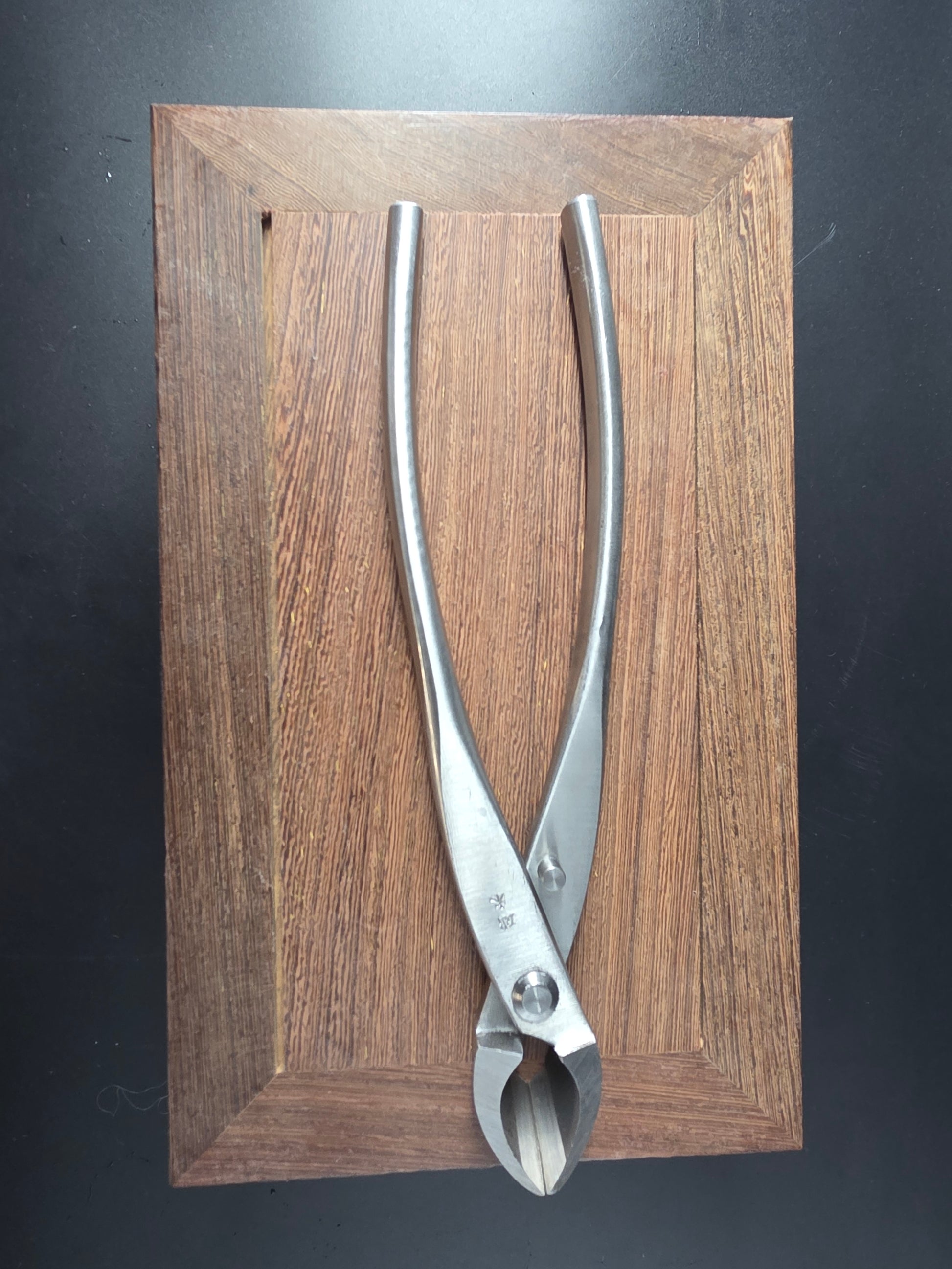 Silver bonsai concave cutter with a curved cutting head and long handles, laid flat on a wooden board