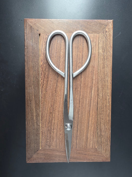 Silver bonsai pruning scissors with loop handles and long narrow pointed blades, laid flat on a wooden board